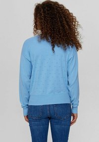 Kvinna med lockigt hår som bär en ljusblå långärmad tröja med små hålmönster och blå jeans, vänd bort mot en enkel bakgrund.