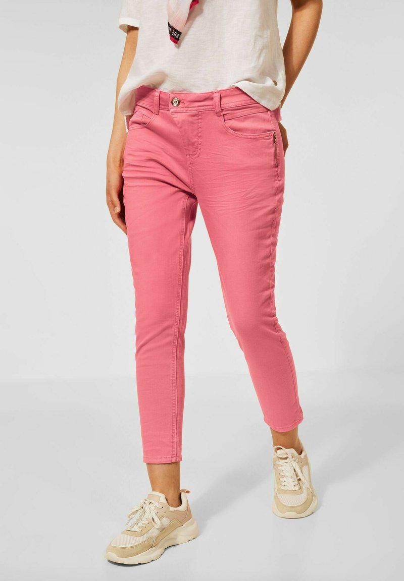 Jean en denim rose avec une coupe slim, cinq poches et une fermeture éclair à l'avant. Portés avec des baskets beiges. Texture lisse et design ajusté.