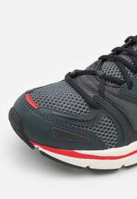 Zapatilla deportiva con parte superior de malla gris, cordones negros, detalles en rojo y una suela de goma negra texturizada. Presenta amortiguación y detalles acolchados.