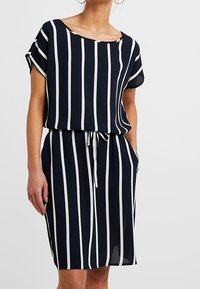 Mekko, joka on tummansininen ja jossa on valkoisia pystysuoria raitoja, lyhyet hihat, pyöreä pääntie ja kiristettävä vyötärö. Materiaali näyttää kevyeltä ja sileältä.