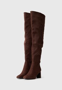 Καφέ γόνατα ψηλά μποτάκια φτιαγμένα από μαλακό σουέτ, με κομψή μύτη, τετράγωνη φτέρνα και ραφιναρισμένες λεπτομέρειες στην κορυφή. Λείος υφής σε όλη την επιφάνεια.