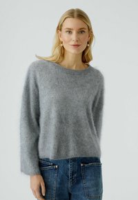 Lichtgrijs, fluffy trui met ronde hals en lange mouwen. Gecombineerd met blauwe spijkerbroek met zichtbare achterzakken.
