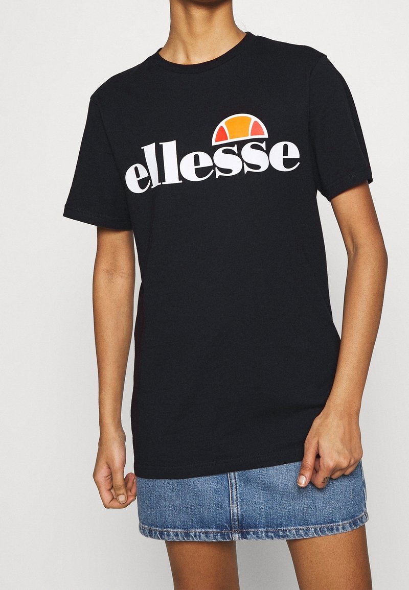 Camiseta de algodón negra con gran logo blanco de "ellesse" y un acento de semicírculo en rojo y naranja, combinada con una falda de mezclilla.