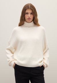 Cremefarbener, gerippter Rollkragenpullover mit lockerem Schnitt, überschnittenen Schultern und langen Ärmeln. Weiche Textur, kombiniert mit schwarzen Hosen für einen Kontrast.