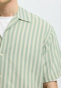 Camisa de manga corta a rayas verde y crema con cuello de pico, que presenta una textura suave y botones en una tela ligera.