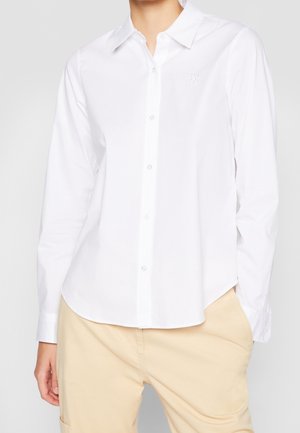 Persoon draagt een nauwsluitend wit overhemd met knopen en lange mouwen en beige broek, met één hand in de zak, tegen een effen achtergrond.