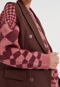 Brown tailored vest with two buttons, layered over a pink patterned knit sweater featuring a diamond design in darker shades of pink.