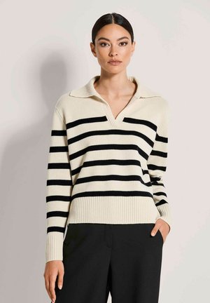 Frau mit dunklen Haaren in einem cremefarbenen Pullover mit schwarzen horizontalen Streifen und schwarzer Hose, stehend mit einer Hand in der Tasche.