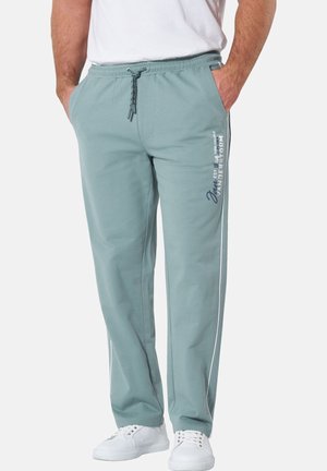 Mann trägt hellgrüne Jogginghosen mit Kordelzug und weißem Aufdruck auf dem linken Oberschenkel, weiße Sneaker und ein weißes T-Shirt.