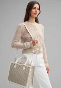 Beżowy sweter dzianinowy z guzikami, biały pasek, białe dżinsy oraz beżowa torba wzorzysta z białymi uchwytami i złotymi akcentami.