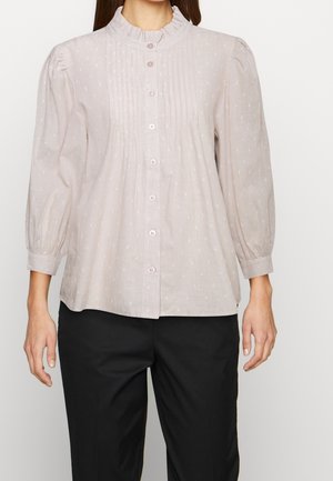 Blouse rose clair avec un col à volants, fermeture à bouton, et de subtils points texturés. Manches longues bouffantes et coupe ample, associée à un pantalon sombre.
