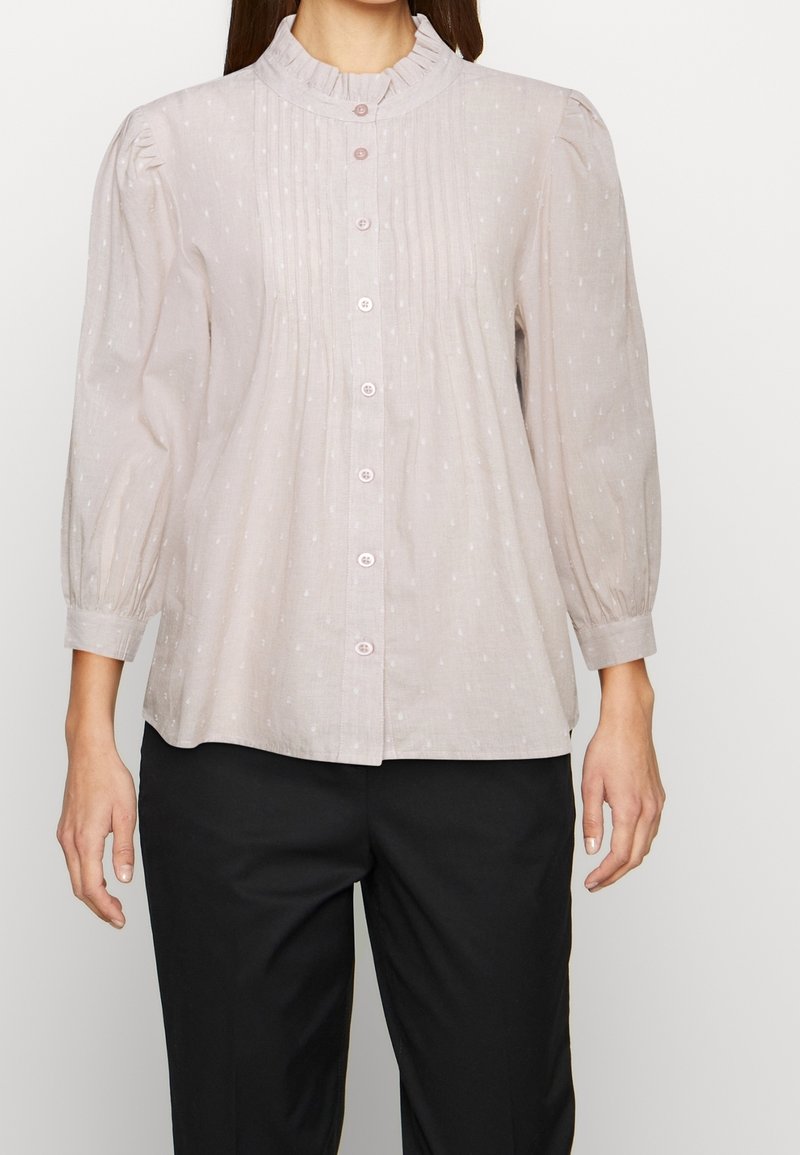 Blusa rosa chiaro con colletto arricciato, profilo a bottoni e discrete macchie testurizzate. Maniche lunghe a sbuffo e vestibilità ampia, abbinata a pantaloni scuri.