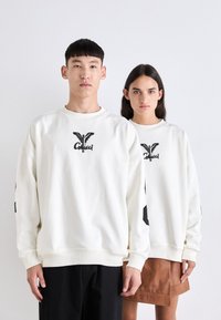 Hvit crewneck hettegenser har en svart grafisk logo på brystet og svarte detaljer på ermene. Stoffet ser glatt og mykt ut.
