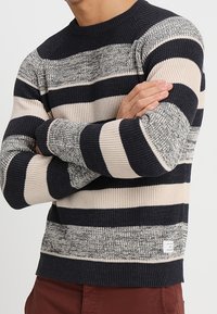 Mężczyzna noszący sweter z prążkowanej dzianiny w szerokie poziome paski w kolorze czarnym, beżowym i szarym, z rękami skrzyżowanymi na piersi.