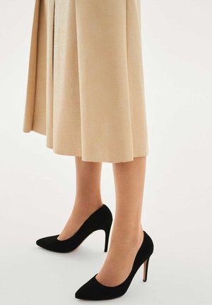 Czarne zamszowe szpilki z ostrym noskiem i cienkim obcasem typu sztylet, zestawione z beżową plisowaną spódnicą o fakturowanym wykończeniu.
