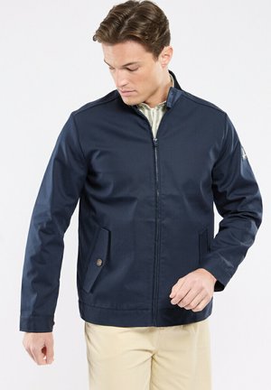Homme portant une veste zippée bleu marine sur une chemise rayée et un pantalon beige, regardant vers le bas et ajustant le poignet de la veste.