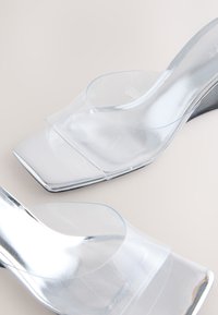 Prozirne akrilne sandale na platformu s kvadratnim vrhom, s glatkim, reflektirajućim završetkom i minimalističkim dizajnom. Prozirni gornji dio s izrezima.