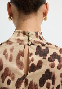 Collo di una donna e parte superiore della schiena con una blusa a stampa animale beige e marrone, chiusa da tre bottoni di perla, indossando piccoli orecchini a cerchio d'oro.