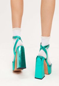 Teal metalliska plattformsskor med tjocka klackar, ankelremmar och en glänsande yta, parat med vita ankelsockor med spetskant.
