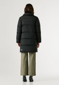 Melna pūkainā jaka ar horizontālu quiltētu dizainu, garām piedurknēm un augstu apkakli, apvienota ar brīvām olīvzaļa biksēm.