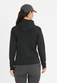 Haut de sport noir avec une surface texturée, une capuche ajustée et des manches longues. Il présente un dos lisse et des accents en maille respirante.