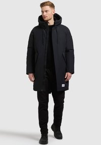 Parka neagră căptușită, cu glugă, închidere cu fermoar și buzunare laterale. Are un finisaj texturat și un patch cu logo în partea din stânga jos.