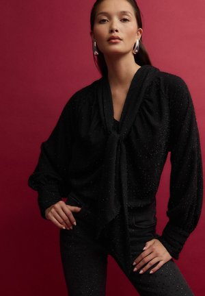 Blouse noire texturée avec un col noué, des manches pleines et des accents scintillants, assortie à un pantalon noir ajusté avec des détails cloutés.
