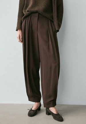 Persona che indossa pantaloni marroni larghi a vita alta con orli risvoltati, abbinati a scarpe marroni con tacco basso e un maglione coordinato.