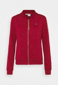 Veste zippée rouge avec col et poignets côtelés, présentant un motif texturé et un accent de logo sur la manche. Détail de fermeture éclair doré.