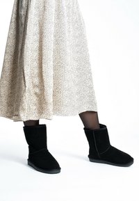 Botas de ante negras hasta el tobillo con punta redonda, forro de borreguito y un puño doblado, combinadas con una falda de beige con manchas a la altura de la rodilla.