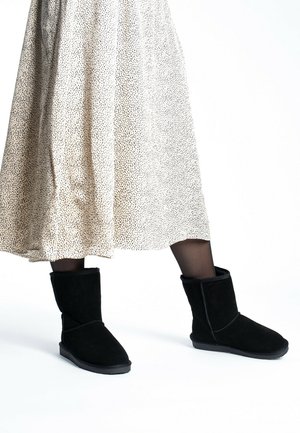 Black suede ankle boots with a rounded toe, shearling lining, and a fold-over cuff, paired with a knee-length beige spotted skirt.
