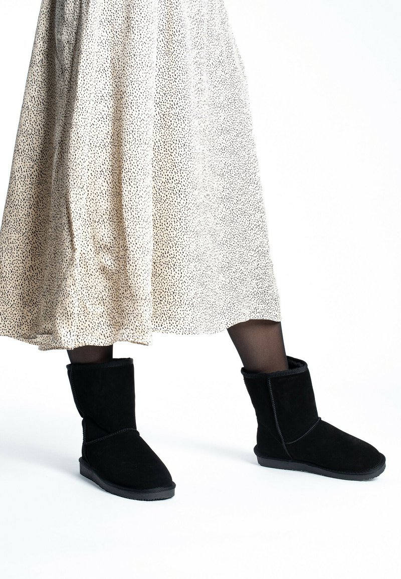 Botas de ante negras hasta el tobillo con punta redonda, forro de borreguito y un puño doblado, combinadas con una falda de beige con manchas a la altura de la rodilla.