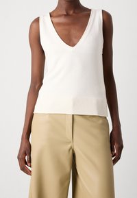 Top senza maniche a V in maglia bianca con orlo a coste. Tessuto liscio, design aderente, abbinato a pantaloni palazzo in finta pelle beige.