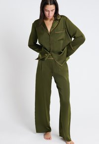 Ensemble de pyjama en satin vert olive comprenant une chemise à boutons avec des passepoils et un pantalon à jambes larges, présentant une texture lisse et brillante.