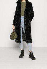 Abrigo de pelo sintético negro con un cuello ancho, suéter verde de canalé, jeans light blue rasgados y botas negras de tobillo, sosteniendo un bolso verde.