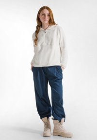 Sudadera de felpa blanca con cuello de media cremallera, combinada con pantalones de mezclilla azules holgados y botas beige con cordones. Textura suave y ajuste relajado.