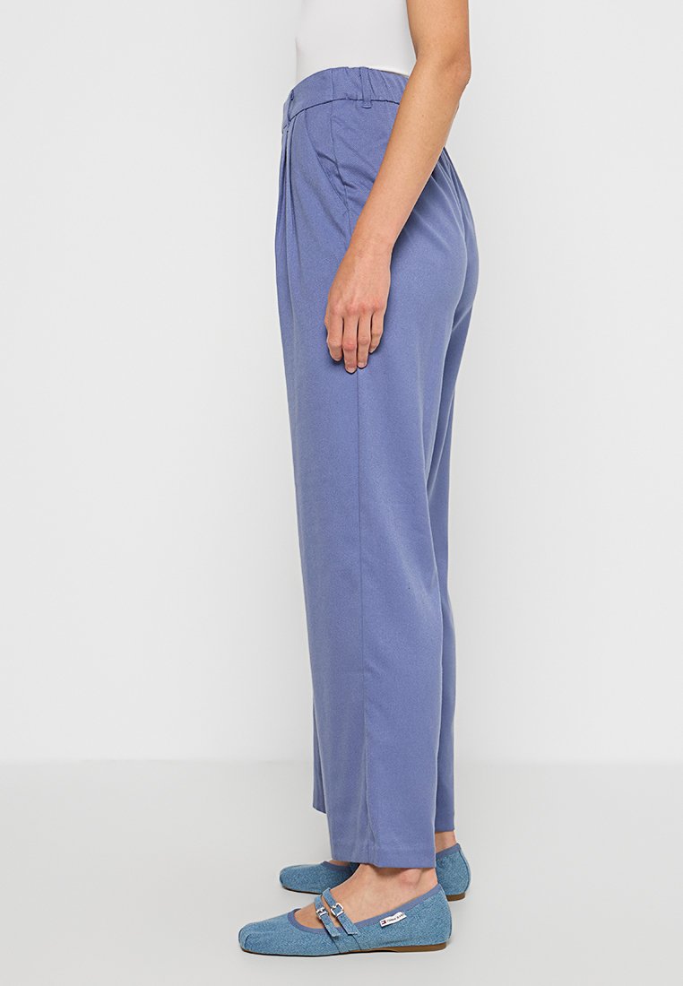 Jasnoniebieskie, zwężane spodnie z elastycznym paskiem i bocznymi zakładkami. Tkanina wydaje się gładka z delikatnym połyskiem. Noszone z jasnoniebieskimi balerinami.