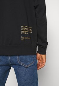 Black long-sleeve sweatshirt with gold printed text on the back, featuring a soft fabric texture and ribbed cuffs, paired with blue denim jeans.