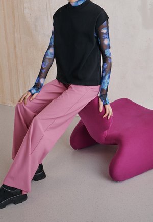 Zwarte mouwloze top gecombineerd met strakke roze broek en gestreepte lange mouwen. Roze abstract gevormde zitplaatsen in een minimalistische binnenruimte.