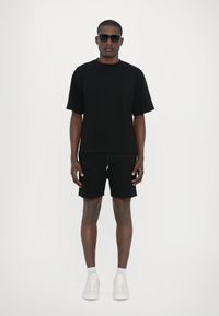 Sort kortærmet t-shirt og shorts, parret med hvide sneakers. Modellen bærer sorte solbriller, med en glat, mat stofstruktur.