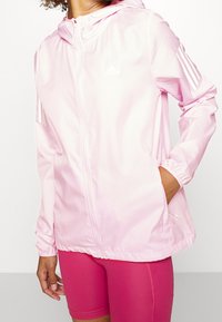 Jasnoróżowa kurtka na zamek, wykonana z lekkiego materiału, z kapturem, elastycznymi mankietami i białym logo Adidas na klatce piersiowej oraz trzema paskami na rękawach.