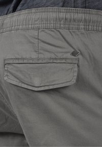 Los pantalones cortos de tela gris presentan un bolsillo trasero con solapa, una textura sutil y una pequeña etiqueta de logo cerca de la costura del bolsillo.