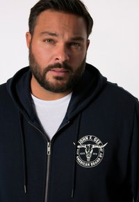 Sweat à capuche zippé bleu marine avec un logo blanc représentant un crâne de taureau. Il possède une capuche avec cordon de serrage et un tissu doux et texturé.