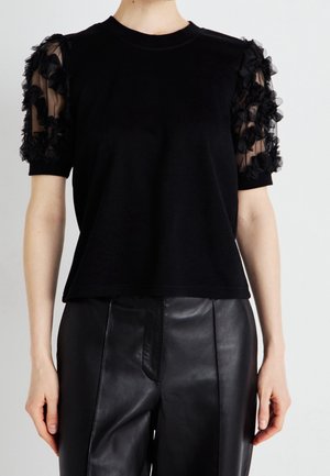 Haut noir à manches courtes avec manches bouffantes transparentes ornées d'appliques florales texturées, porté avec un pantalon en cuir noir.