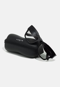 Schwarze Kunststoff-Sonnenbrille mit geometrischem Design, die auf einem schwarzen Reißverschlussetui liegt, auf dem der Markenname "vogue" deutlich zu sehen ist.
