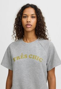 Pelēks kokvilnas īsām piedurknēm svārku tops ar tekstu "TRÈS CHIC" zeltā, ar brīvu piegriezumu un raglanu piedurknēm.