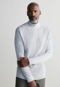 Hellgrauer Pullover mit Reißverschluss am Hals, der ein strukturiertes Strickmuster und gerippte Bündchen aufweist. Der glatte Stoff erstreckt sich bis zum Taillenbund.