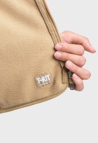 Sac en polaire beige avec une surface texturée, doté d'une fermeture éclair, d'un patch logo et d'une main tenant la tirette de la fermeture.