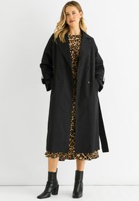 Black double-breasted trench coat with large lapels, side pockets, and a tie belt, layered over a leopard print dress and ankle boots.
