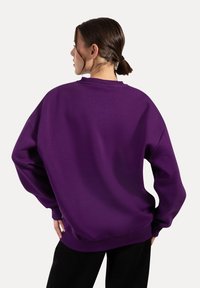 Sudadera morada de gran tamaño con cuello redondo ribeteado y puños, que presenta una textura suave y un corte relajado, vista desde la parte de atrás.
